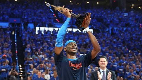 Basketball player in Thunder jersey raising an award above their head before a crowd in a blue-lit arena