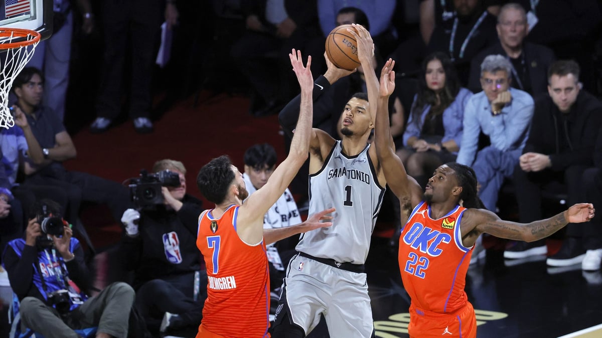 Basketball player in white jersey shooting over two defenders in orange jerseys during an NBA game with crowd in background